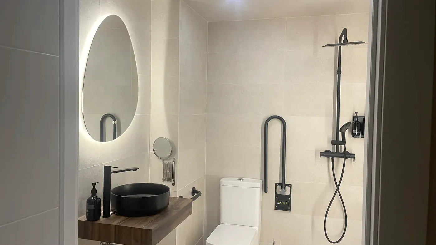 Baño con lavabo sobre repisa de madera, inodoro con barra abatible y ducha a ras de suelo