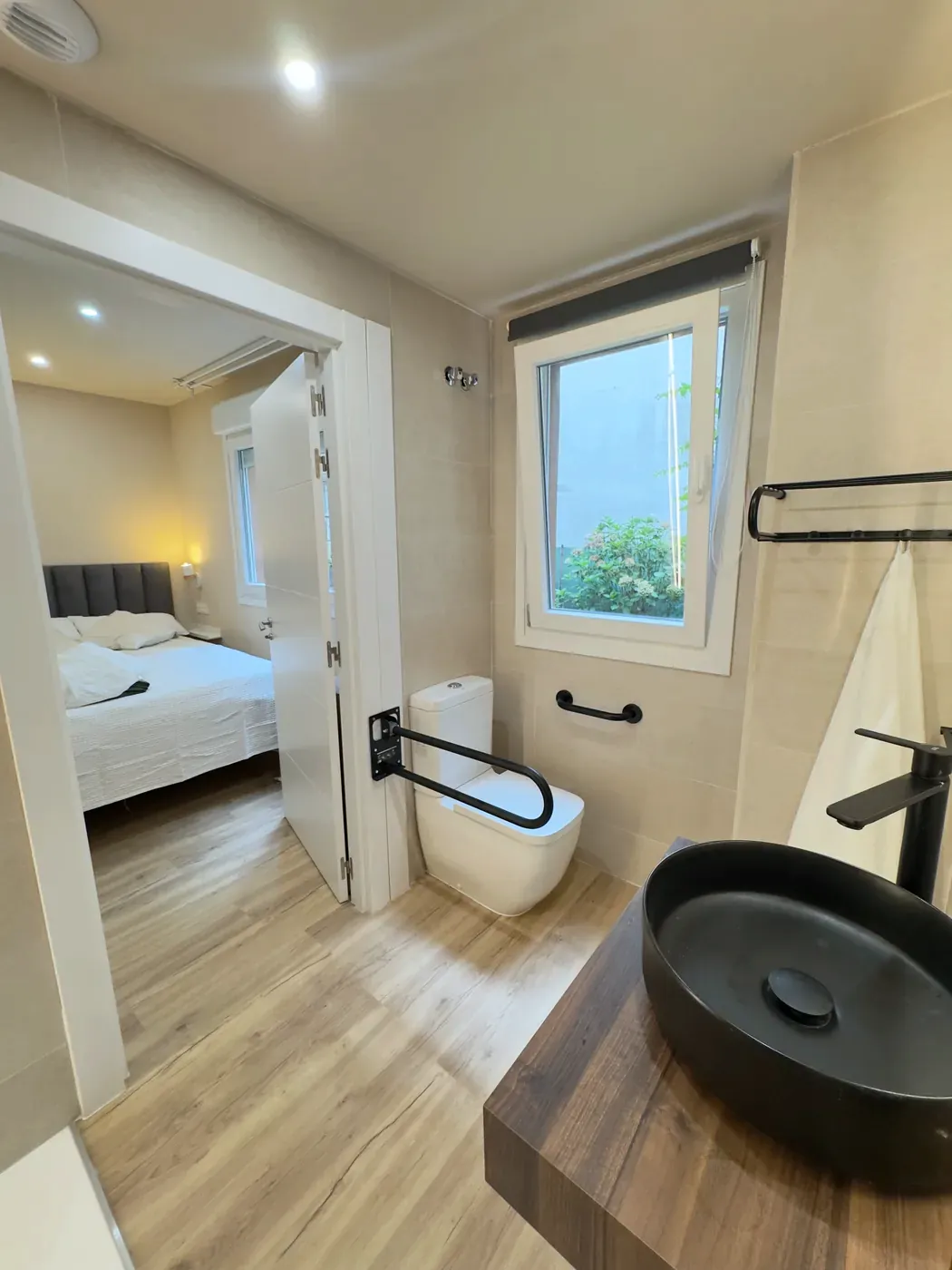 Baño con barras de apoyo junto al inodoro, visto desde el dormitorio